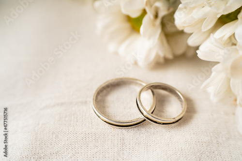 Pair of wedding rings on a white surface with beautiful white flowers. Commitment and love concept. Copy Space, differential focus.