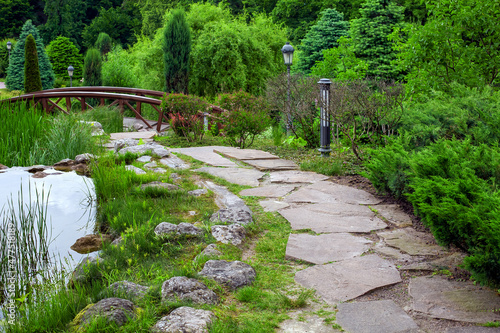 natural stone path along pond with water and reeds among green backyard plants with evergreen bushes and pine trees landscape design of eco friendly summer park with lantern, nobody.