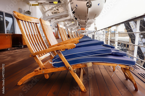 Deckchairs, with blue foldec cushions, lined up on a cruise line on a cloudy port day. Liferafts can be seen hanging above the deck. With space for text.
