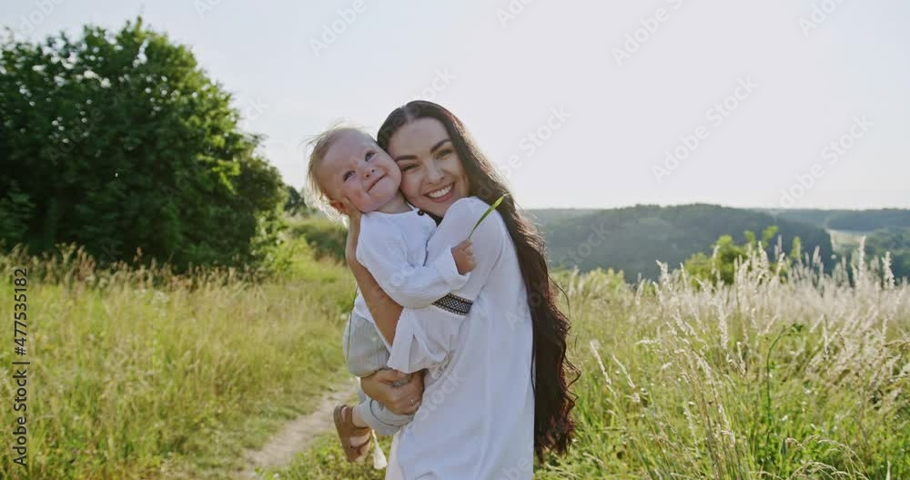Beautiful family portrait of a loving mother with her baby both smiling happily at the camera outdoors in a rural landscape as she hugs the infant to her cheek Beautiful family portrait of a loving mother with her baby both smiling happily at the camera outdoors in a rural landscape as she hugs the infant to her cheek