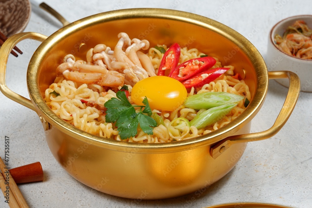 Serving a Korean style instant noodle, Ramyeon or Ramyun with spicy flavour topped with egg yolk, chilli, vegetables and kimchi in a traditional Korean noodle pot. Serving a Korean style instant noodle, Ramyeon or Ramyun with spicy flavour topped with egg yolk, chilli, vegetables and kimchi in a traditional Korean noodle pot.