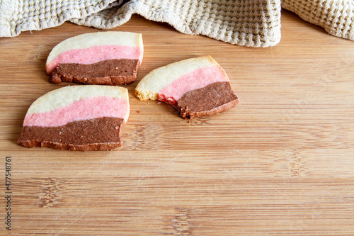 Delicious white, pink and chocolate cookies and one with a bite taken out; Fresh homemade Neapolitan cookies on a wooden cutting board
