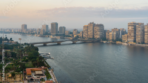 View of the Nile in Cairo from a height. The bridge passes over the blue river. On the banks - high-rise buildings, city houses, green vegetation. Clouds in the pinkish evening sky. Egypt