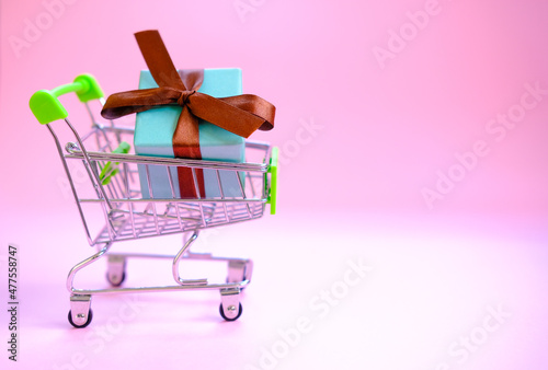 A present box with a ribbon in a shopping trolley cart on pink background . Concept - buying a gift ,online and sale. Christmas, valentines ,women day .Copy space, mockup.