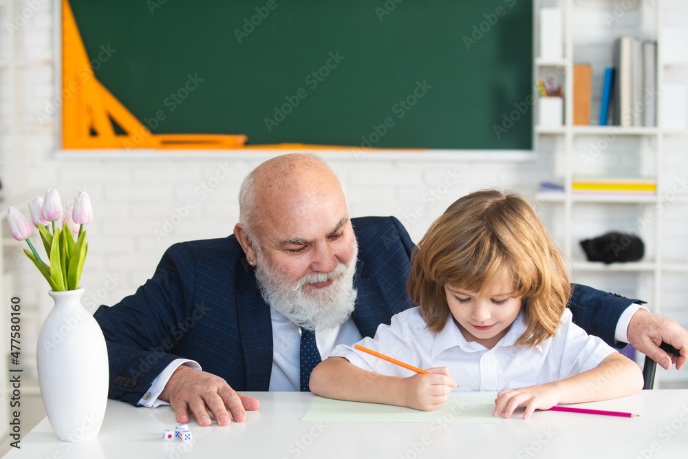 Senior teacher or grandfather and school boy pupil in classroom at school. Elementary pupil reading and writing with teacher in classroom. Senior teacher or grandfather and school boy pupil in classroom at school. Elementary pupil reading and writing with teacher in classroom.