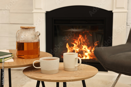 Fototapeta Cups of hot drink and teapot on wooden tables near decorative fireplace in room