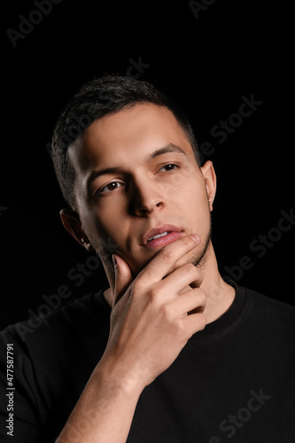 Thoughtful young man pondering deeply with hand on chin, expressing contemplation and problem solving on a dark background