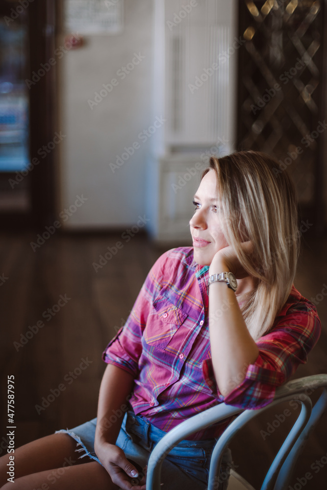 Adult shining blonde woman with face looking away, sitting on chair in room leaning on chair back with left hand wearing checkered shirt tied in belly area and jeans skirt in daytime.