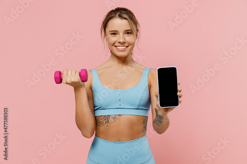 Young strong sporty fitness trainer instructor woman wear blue tracksuit spend time in home gym hold female dumbbells blank screen mobile phone isolated on plain pink background Workout sport concept