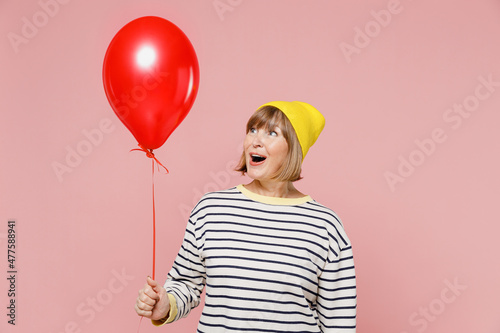 Elderly amazed woman 50s in striped shirt yellow hat celebrating birthday holiday party and hold colorful air inflated helium balloon isolated on plain pastel light pink background studio portrait photo