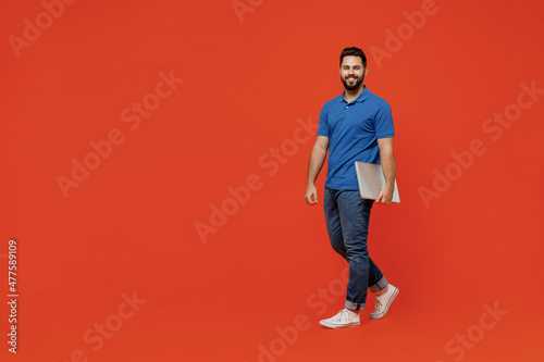 Full body young smiling confident happy caucasian man 20s wear basic blue t-shirt looking camera hold closed laptop pc computer walk go isolated on plain orange background People lifestyle concept
