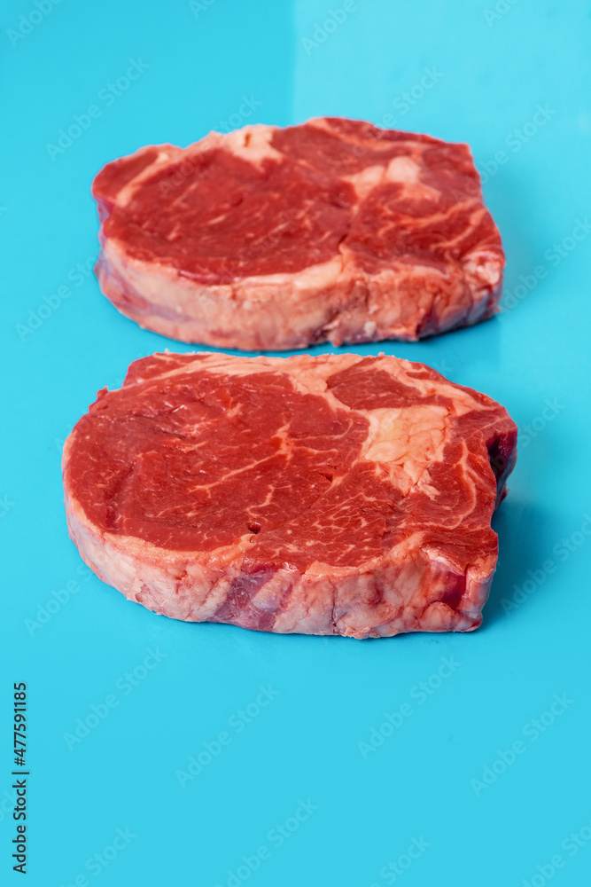 Two fresh juicy rib eye steak on blue surface. Top quality fresh meat. Butcher craft product and meat industry. Rich red color and marbling. Premium cut. Two fresh juicy rib eye steak on blue surface. Top quality fresh meat. Butcher craft product and meat industry. Rich red color and marbling. Premium cut.