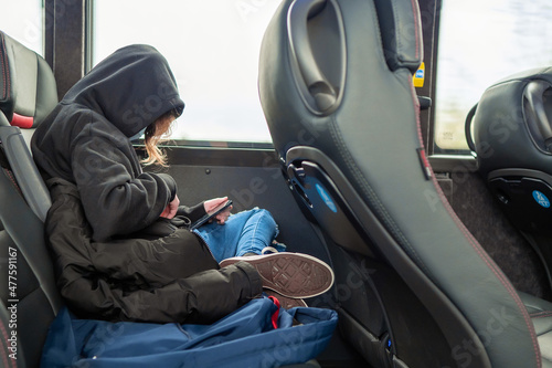 Young teenager girl on a trip in a bus watching movie on her smart phone. Kids attached to modern technology concept. The model wears blue jeans and dark hoodie and face mask.