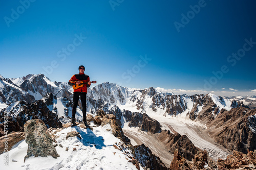 Young man trekker playing guitar and singing song on top of Pik Uchitel peak . Ala Archa Alpine National Park Landscape near Bishkek, Tian Shan Mountain Range, Kyrgyzstan, Central Asia.