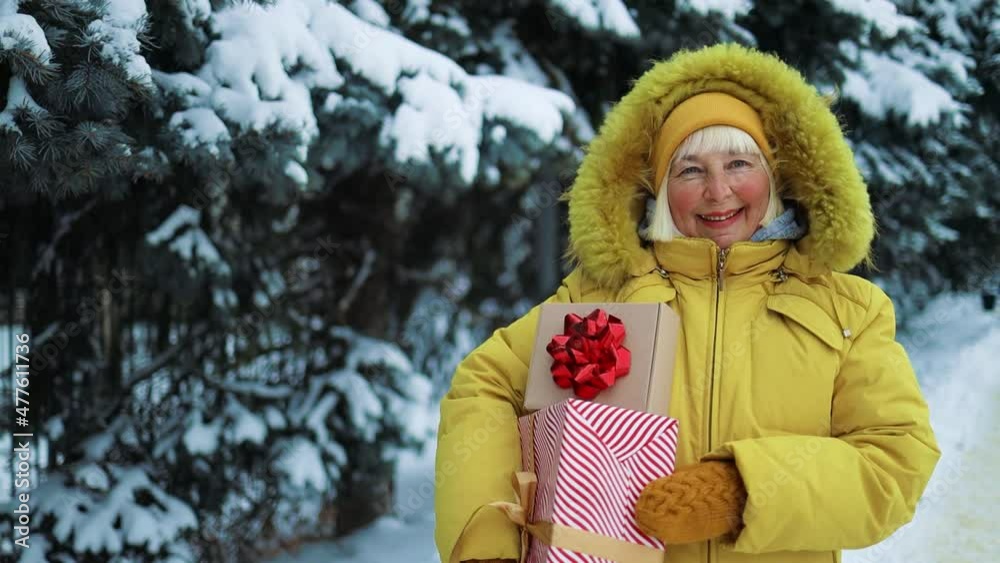 Positive Caucasian 50s woman in yellow clothes hands holding gift boxes and waving hand on christmas shopping outdoors in city. Positive Caucasian 50s woman in yellow clothes hands holding gift boxes and waving hand on christmas shopping outdoors in city.