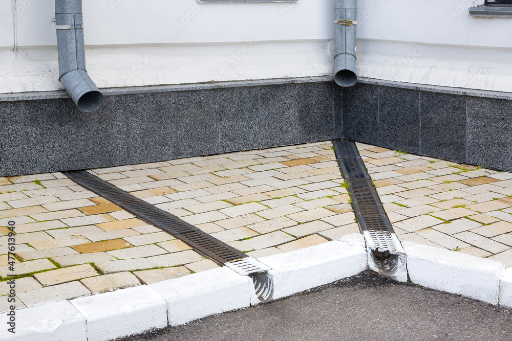Obraz premium facade of a building wall with a downspout pipe and a concrete dish canal with iron grid on a stone slab sidewalk to drain rainwater near asphalt road city street, nobody.