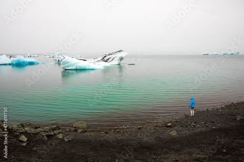 A person in a blue jacket standing on the black coastline in misty weather looking at blue icebergs in the turquoise  icy lake, emerging from the fog and from glaciers behind, Iceland photo