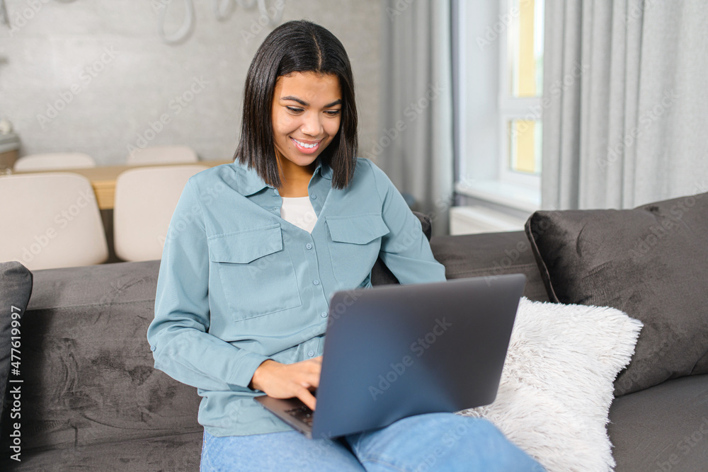 Naklejka premium Multiracial young woman spending home leisure with a laptop. Smiling girl sitting at the cozy sofa and looking at laptop screen. Web browsing, watching TV series, shopping in network concept