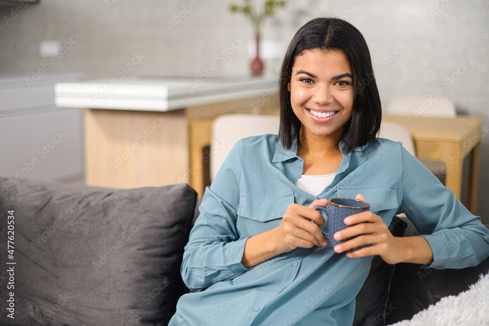 Charming teen lady holding mug of hot drink, looking at the camera, daydreaming and holding coffee, sitting on the couch in a cozy living room, enjoying free time Charming teen lady holding mug of hot drink, looking at the camera, daydreaming and holding coffee, sitting on the couch in a cozy living room, enjoying free time
