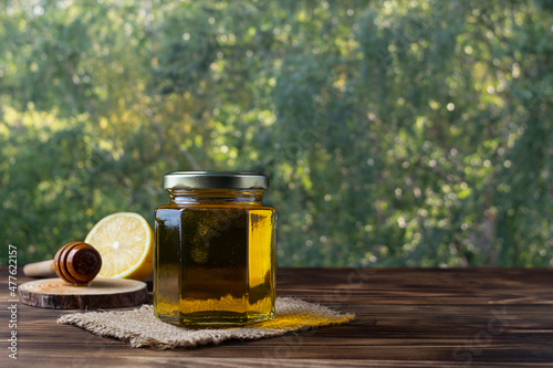 Transparent golden honey in a glass jar on a canvas napkin. Half a lemon and a spoon for honey in the background. Green background with bokeh. Dark brown wooden table. Place for text