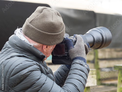 A man in winter clothing holds a professional camera with a long telephoto lens wearing gloves and a hat