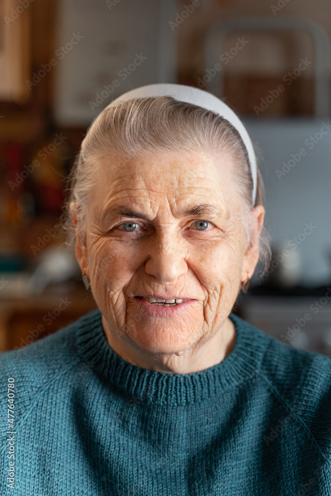 The face of a happy elderly woman of the 80s. He smiles and looks at the camera. Portrait of an old lady with gray hair in a knitted green jacket. There is a blurry image of the kitchen. The face of a happy elderly woman of the 80s. He smiles and looks at the camera. Portrait of an old lady with gray hair in a knitted green jacket. There is a blurry image of the kitchen.