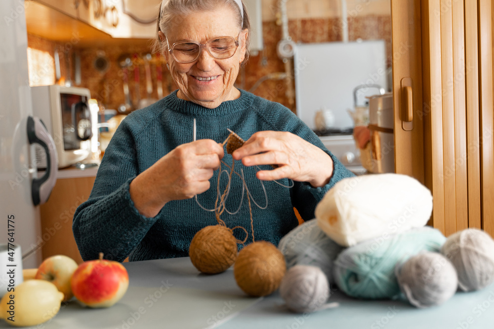 Fototapeta premium Happy elderly woman is engaged in her favorite hobby, knitting while sitting in the kitchen. Glasses for vision. There are threads on the table, a cup of tea, apples and a lemon.