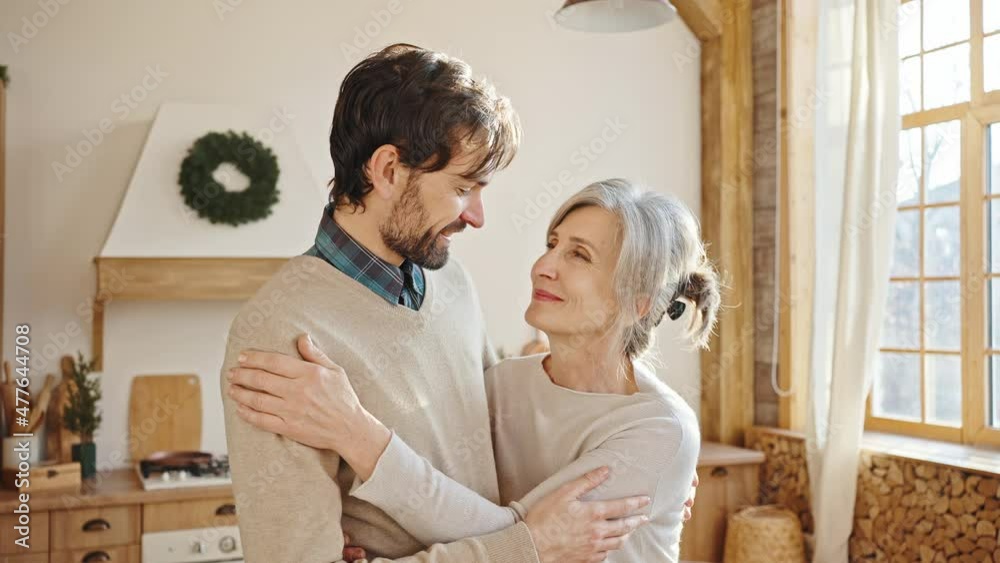 Tracking shot portrait of loving adult son embracing his senior grey-haired mother, smiling together to camera at home Tracking shot portrait of loving adult son embracing his senior grey-haired mother, smiling together to camera at home
