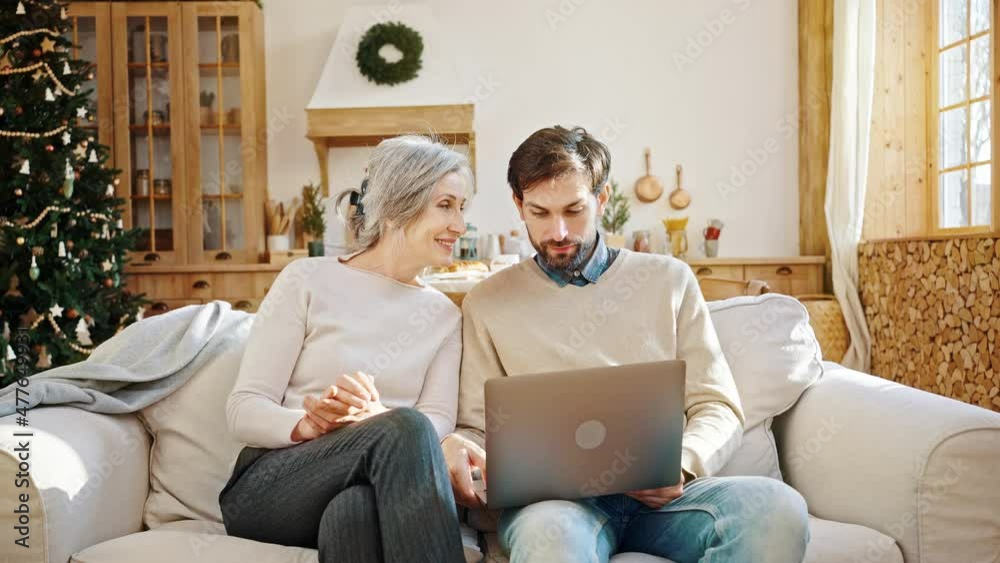 Young positive man showing how to work on laptop to his curious elderly mother, sitting together on sofa at home Young positive man showing how to work on laptop to his curious elderly mother, sitting together on sofa at home