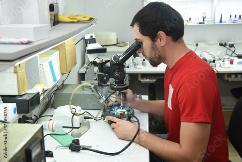 A young man in the workshop examines the microcircuit of an electrical device through a microscope, performing electrical soldering of equipment