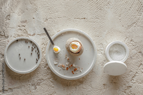 One boiled egg in stoneware egg cup with egg shell broken pieces in concrete tray, salt flakes and pepper in white concrete bowls on rough textured clay background. Healthy breakfast concept.