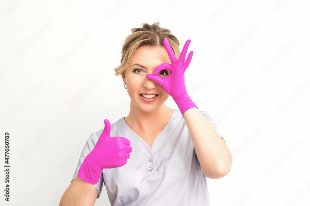 Smiling doctor oculist caucasian woman wearing pink rubber gloves in uniform showing ok sign covering the eye and thumb up gesture against a white background Smiling doctor oculist caucasian woman wearing pink rubber gloves in uniform showing ok sign covering the eye and thumb up gesture against a white background