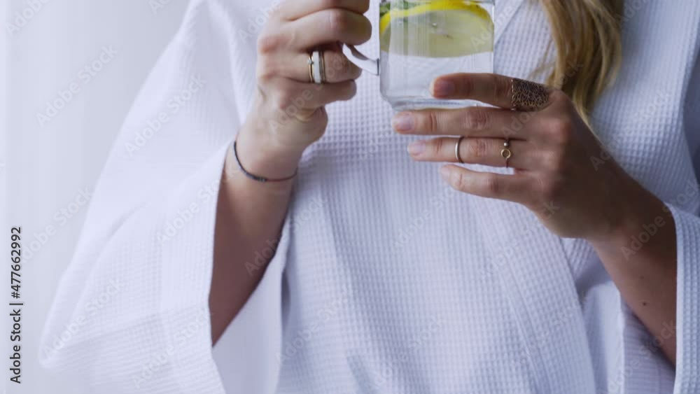 Unrecognizable woman in white bathrobe drinks delicious infused beverage with lemon and mint in light hotel room in morning close view. morning routine, healthy Eating. diet Concept. Unrecognizable woman in white bathrobe drinks delicious infused beverage with lemon and mint in light hotel room in morning close view. morning routine, healthy Eating. diet Concept.
