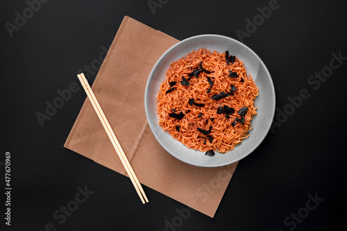 Korean-style instant noodles in a white bowl laid out on brown paper with chopsticks on the side, photographed from above and against a black background. Used in food and health applications