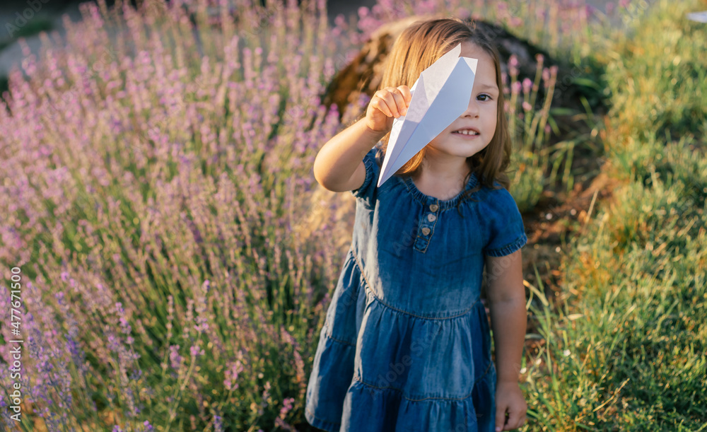 Little girl 3-4 with dark hair in denim dress in sun launches paper plane among large bushes of lilac lavender