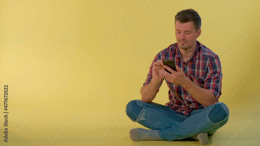 Handsome boy sitting on the floor and chatting with friends on smartphone. He having rest on yellow background. Handsome boy sitting on the floor and chatting with friends on smartphone. He having rest on yellow background.