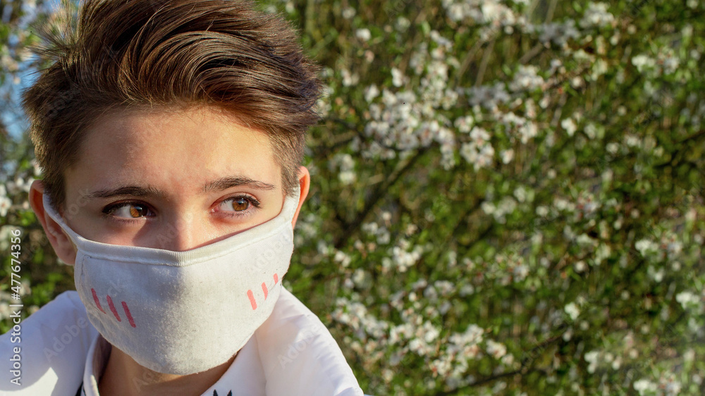 Close-up of teenager face in protective medical tissue mask. Short brown hair. Protection against influenza, virus and pollen plant. Allergy to pollen in spring. On background of blooming cherry tree. Close-up of teenager face in protective medical tissue mask. Short brown hair. Protection against influenza, virus and pollen plant. Allergy to pollen in spring. On background of blooming cherry tree.