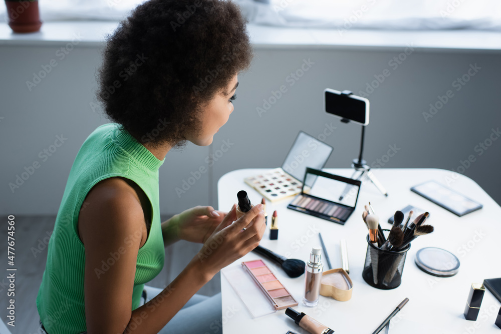 Side view of african american blogger holding face foundation near cosmetics and smartphone on table.