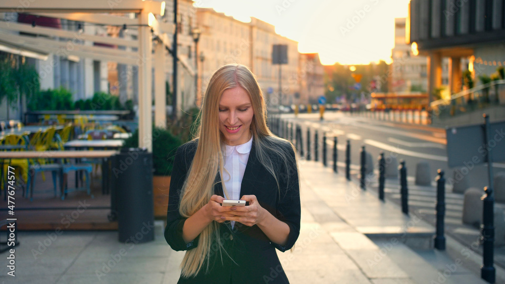 Elegant blond woman in suit walking on street and browsing smartphone with smile against urban background. Elegant blond woman in suit walking on street and browsing smartphone with smile against urban background.