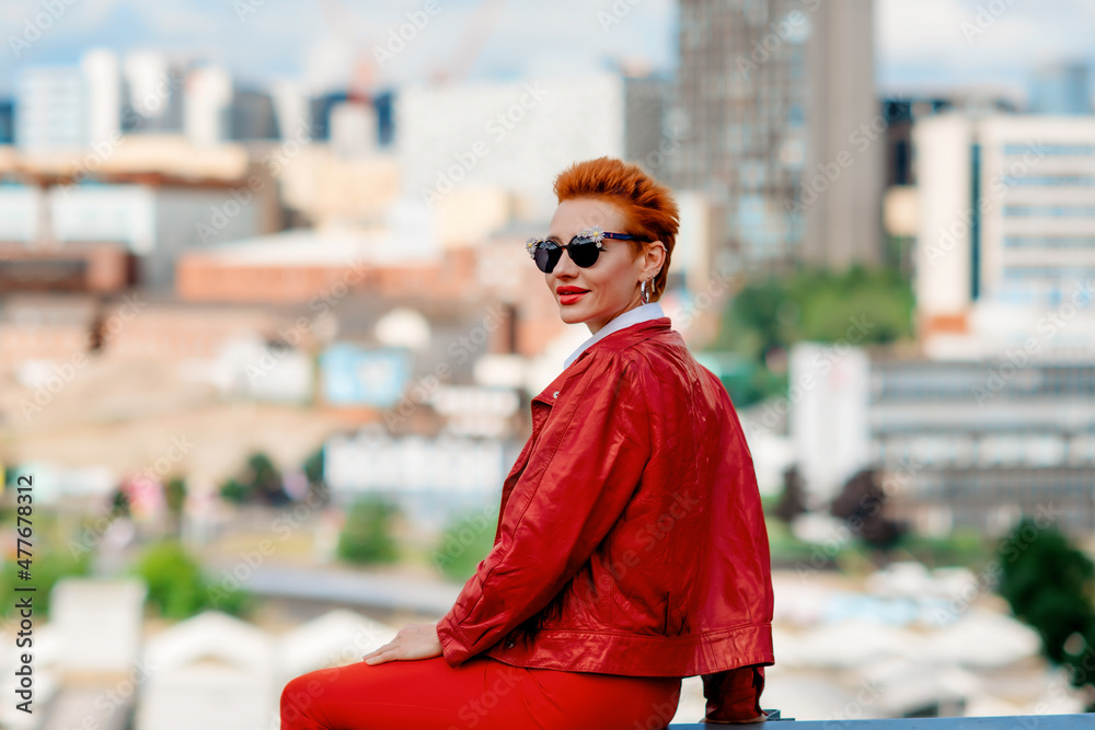 Obraz premium a red-hair woman in a red trousers and coat sitting in front of the city on an summer day