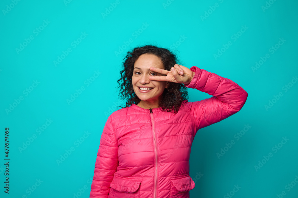 Emotional portrait of beautiful curly-haired woman smiling with cheerful toothy smile looking at camera through a peace sign she shows gesturing her hand, isolated on blue background with copy space Emotional portrait of beautiful curly-haired woman smiling with cheerful toothy smile looking at camera through a peace sign she shows gesturing her hand, isolated on blue background with copy space