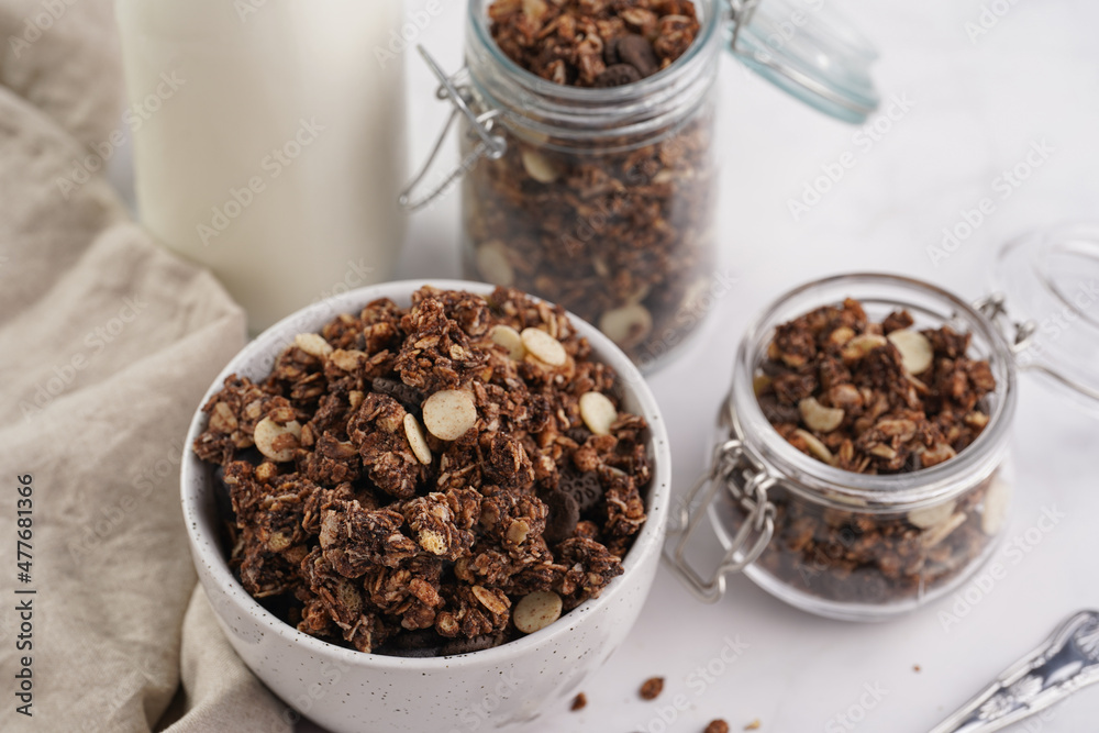 Breakfast muesli cereal with chocolate chips and nuts in several jars and bowl, bottle of milk, white surface Breakfast muesli cereal with chocolate chips and nuts in several jars and bowl, bottle of milk, white surface