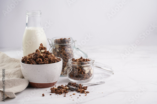 Breakfast muesli cereal with chocolate chips and nuts in several jars and bowl, bottle of milk, white surface