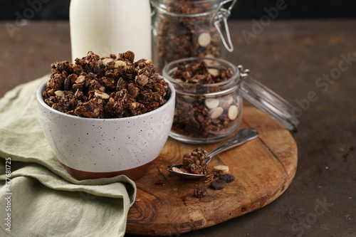 Breakfast muesli cereal with chocolate chips and nuts in several jars and bowl, bottle of milk, brown wooden surface