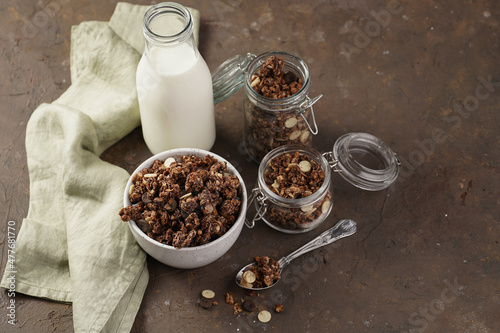 Breakfast muesli cereal with chocolate chips and nuts in several jars and bowl, bottle of milk, brown wooden surface
