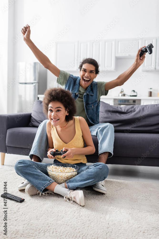 KYIV, UKRAINE - NOVEMBER 9, 2021: Excited african american man holding joystick near girlfriend and popcorn at home. KYIV, UKRAINE - NOVEMBER 9, 2021: Excited african american man holding joystick near girlfriend and popcorn at home.