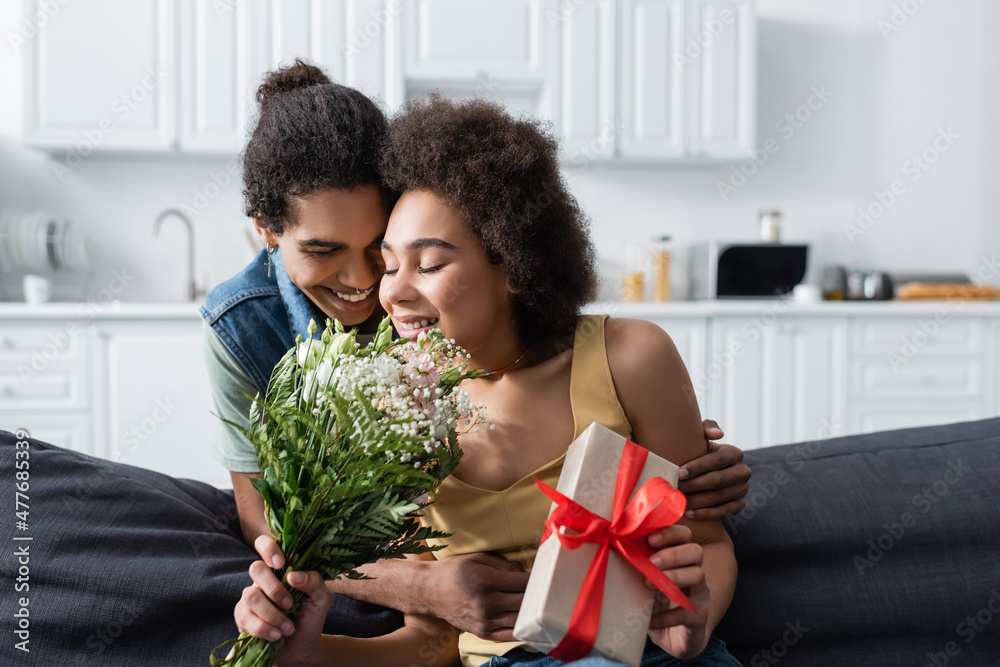 Young african american man hugging smiling girlfriend with bouquet and present at home. Young african american man hugging smiling girlfriend with bouquet and present at home.