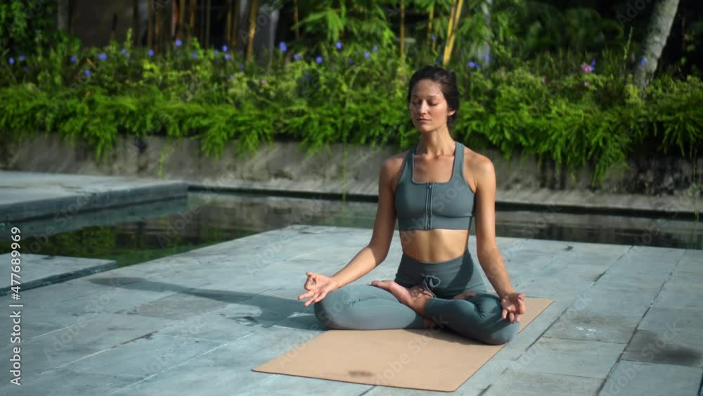 European brunette sports girl makes yoga in nature with a green rainforest on a lit background by the pool on a mat and breathes deeply.