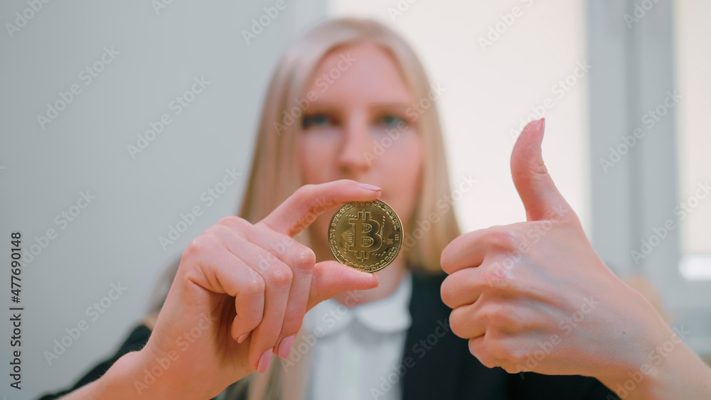 Smiling cheerful blond woman in office suit sitting at workplace with computer and showing bitcoin in hand doing thumbs up gesture and looking at camera Smiling cheerful blond woman in office suit sitting at workplace with computer and showing bitcoin in hand doing thumbs up gesture and looking at camera
