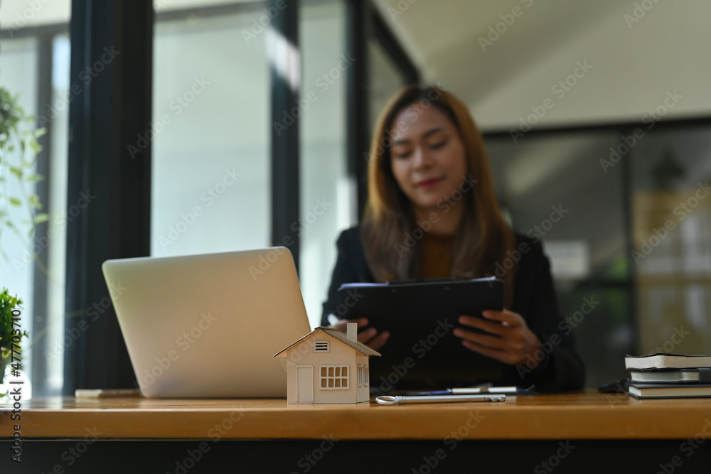 Photo of a young businesswoman holding a clipboard while sitting at the wooden working desk surrounded by a computer laptop, house model and office equipment. Photo of a young businesswoman holding a clipboard while sitting at the wooden working desk surrounded by a computer laptop, house model and office equipment.
